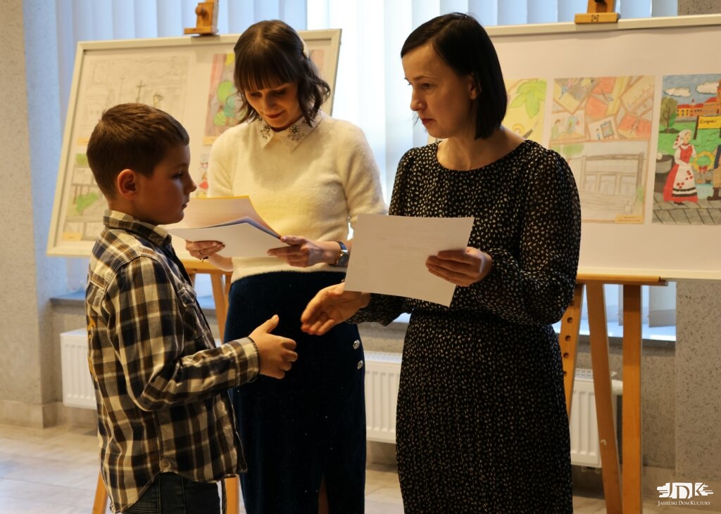 od lewej: chłopiec odbierający dyplom z rąk pani dyrektor JDK, a w tle pani plastyczka JDK