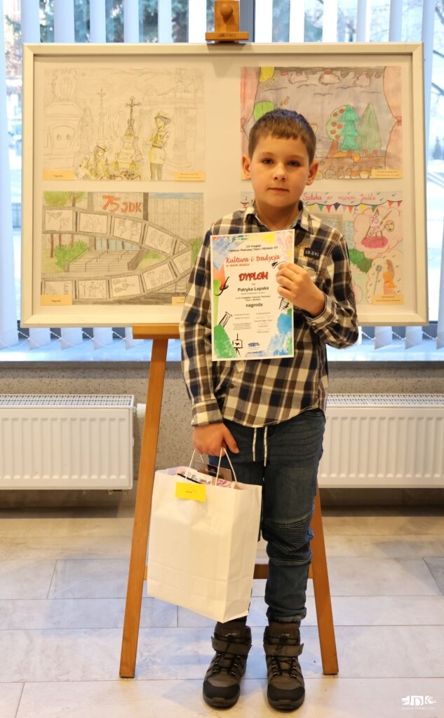 chłopiec w białej koszuli w czarną kratę pozuje do zdjęcia trzymając w ręku dyplom oraz torbę z upominkiem
