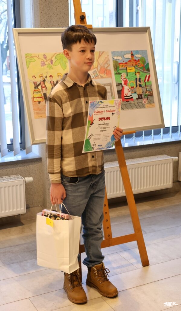 chłopiec w koszuli w dużą kratę, jeansach i butach zimowych trzyma w jednej ręce dyplom, w drugiej torbę z nagrodą
