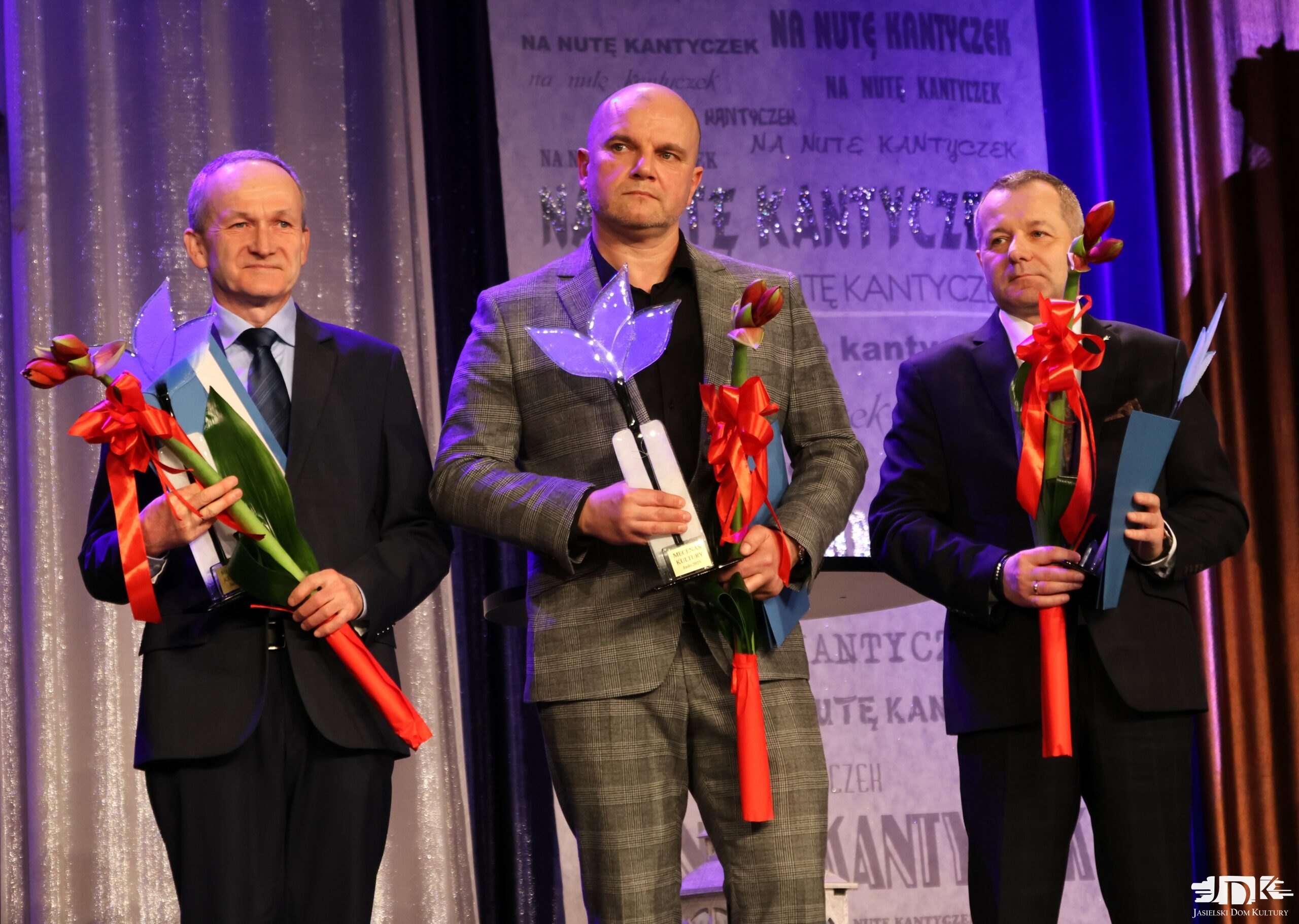 troje mecenasów kultury: od lewej: mężczyzna w czarnym garniturze, na środku mężczyzna w garniturze w kratkę, po prawej mężczyzna w czarnym garniturze. Wszyscy trzymają kwiaty kultury - jasnofioletowe statuetki oraz amarylisy