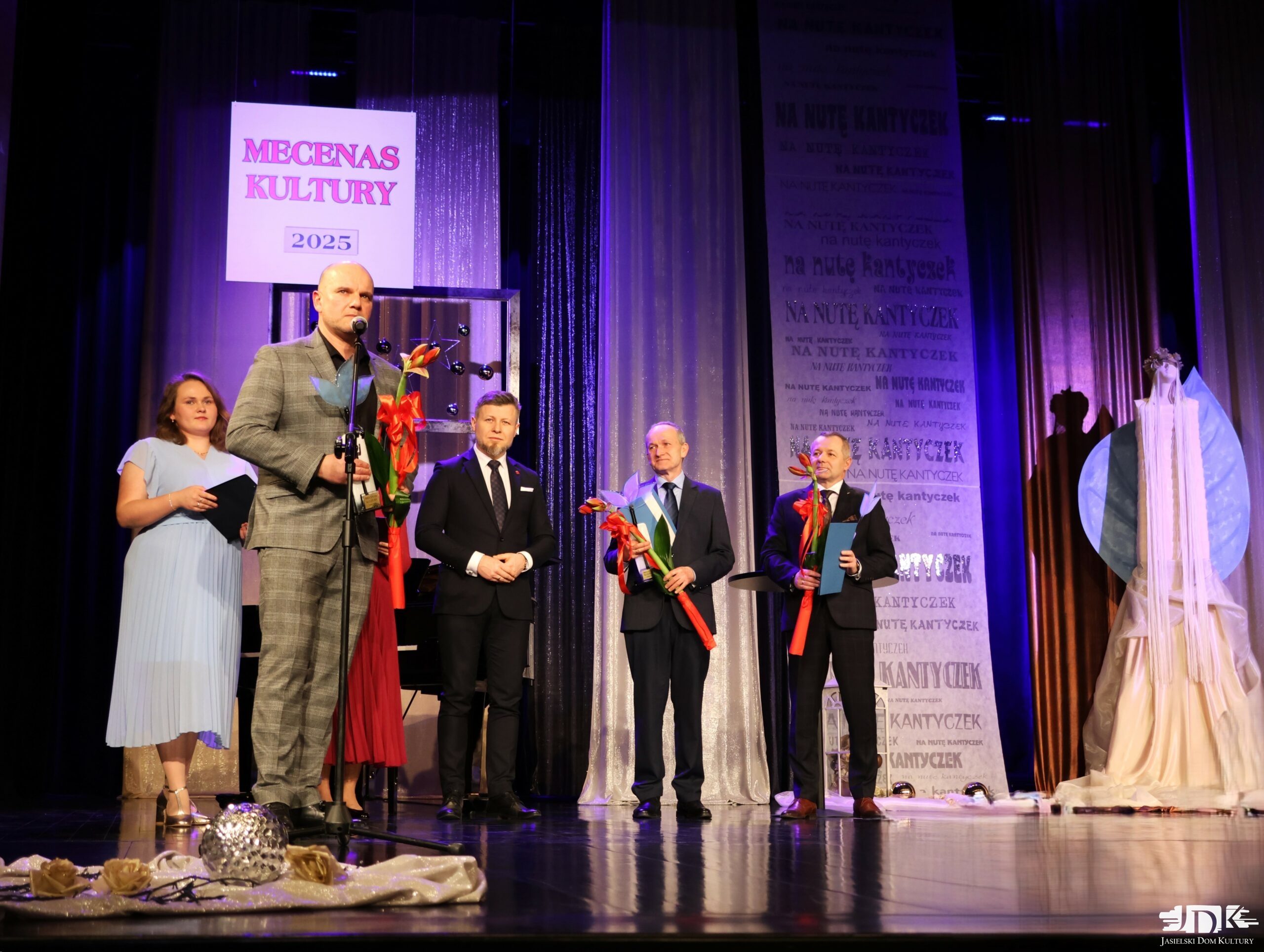 przemawiający mecenas kultury - mężczyzna w garniturze w kratkę, trzymający statuetkę oraz czerwony amarylis