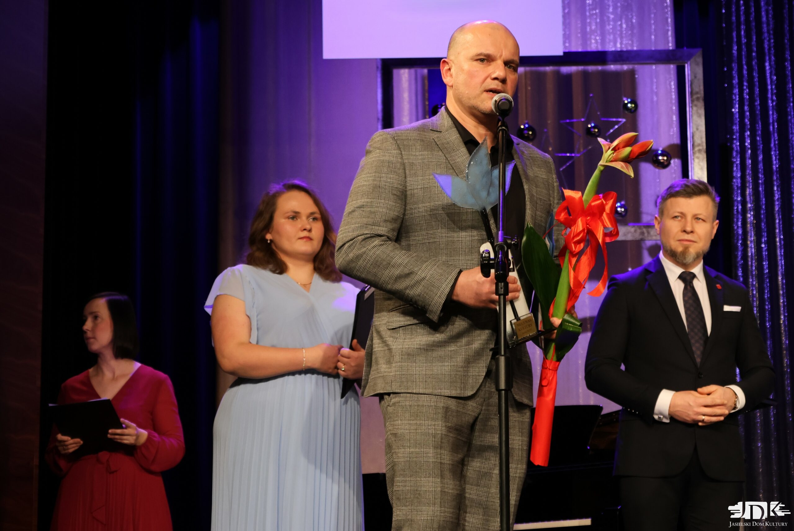 przemawiający mecenas kultury - mężczyzna w garniturze w kratkę, trzymający statuetkę oraz czerwony amarylis