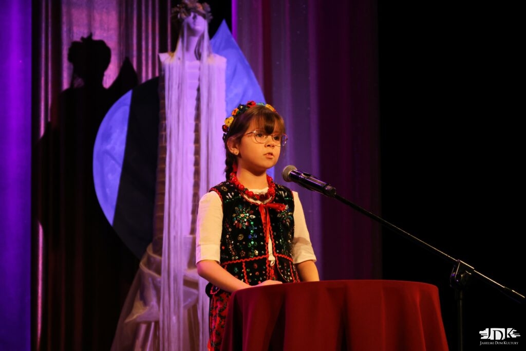 dziewczynka w okularach i stroju ludowym śpiewa zza stołu, za nią dekoracja w postaci anioła