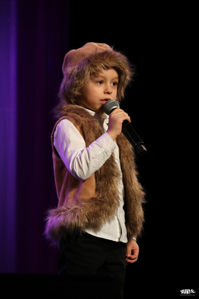 chłopiec z mikrofonem w stroju: brązowa czapka z futrem oraz kożuch obszyty futerkiem