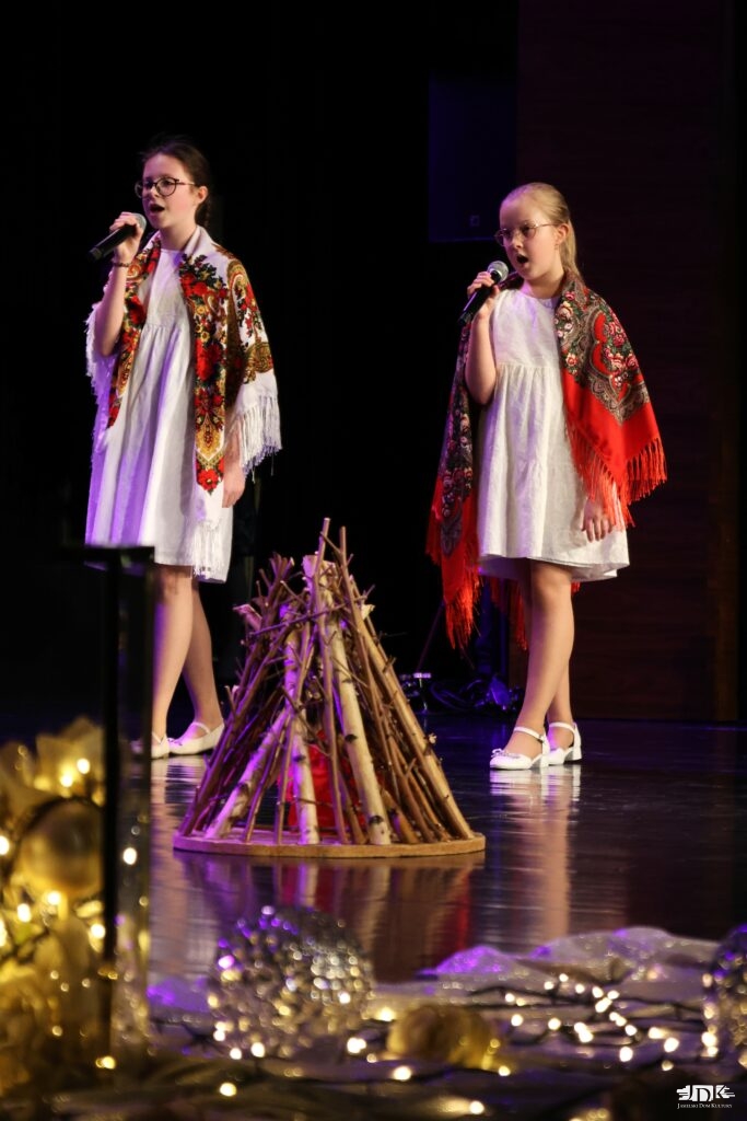 dziewczynki z mikrofonami w białych sukienkach z chustami na ramionach, przed nimi prowizoryczne ognisko