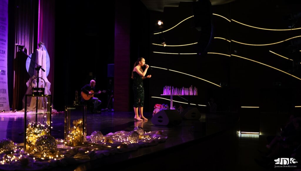 dziewczyna na środku sceny, za nią akompaniator na gitarze akustycznej. Z przodu na proscenium dekoracja świetlna