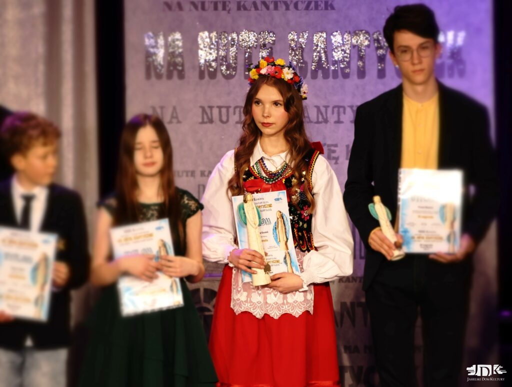 dziewczyna w ludowym stroju z kwiatami we włosach, trzymająca dyplom oraz anioła - nagrodę