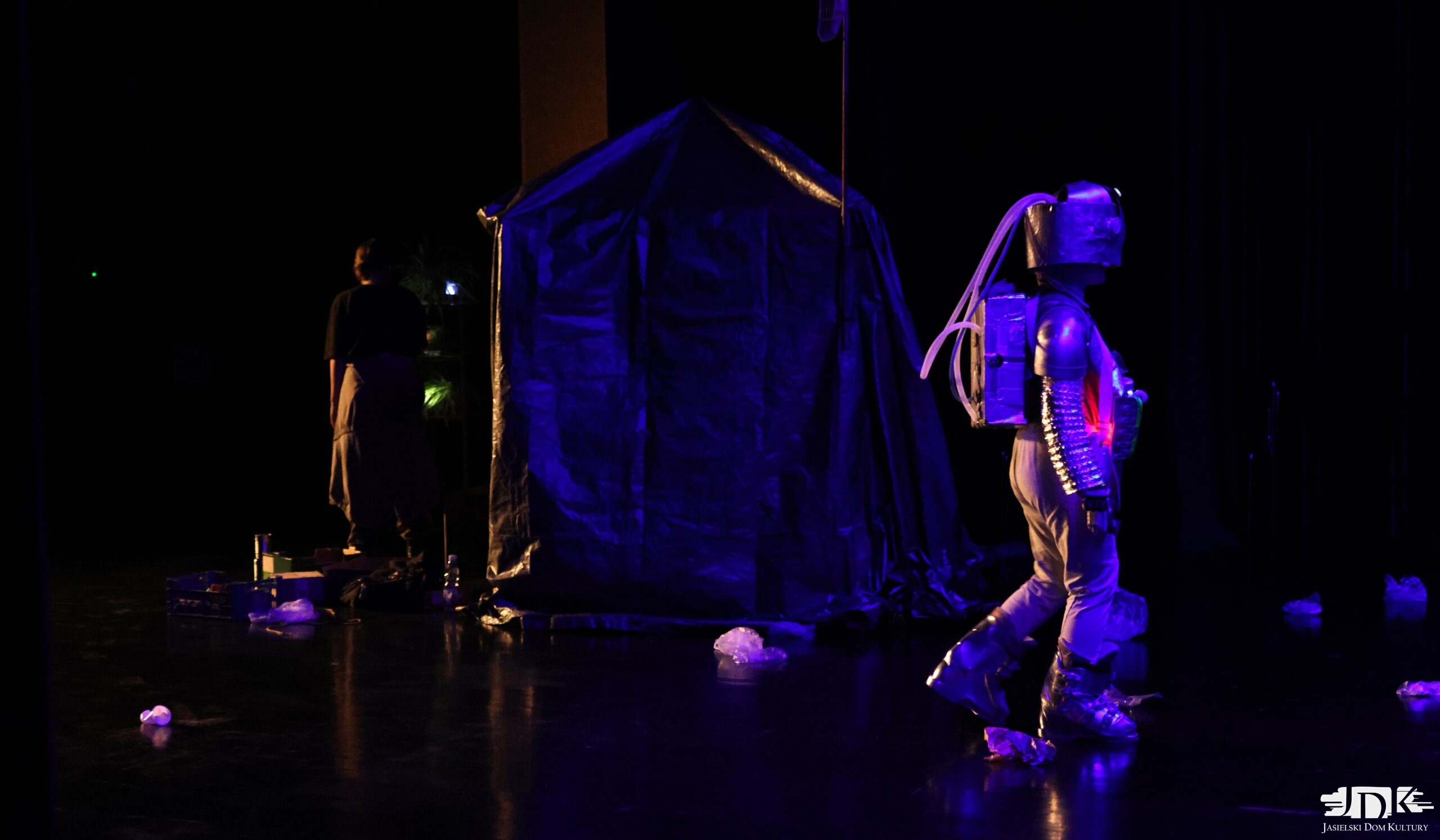 dziewczyna przebrana za robota - srebrny kostium z migającymi światełkami, a w tle prowizoryczny szałas z plandeką, wszystko skąpane niebieskim światłem