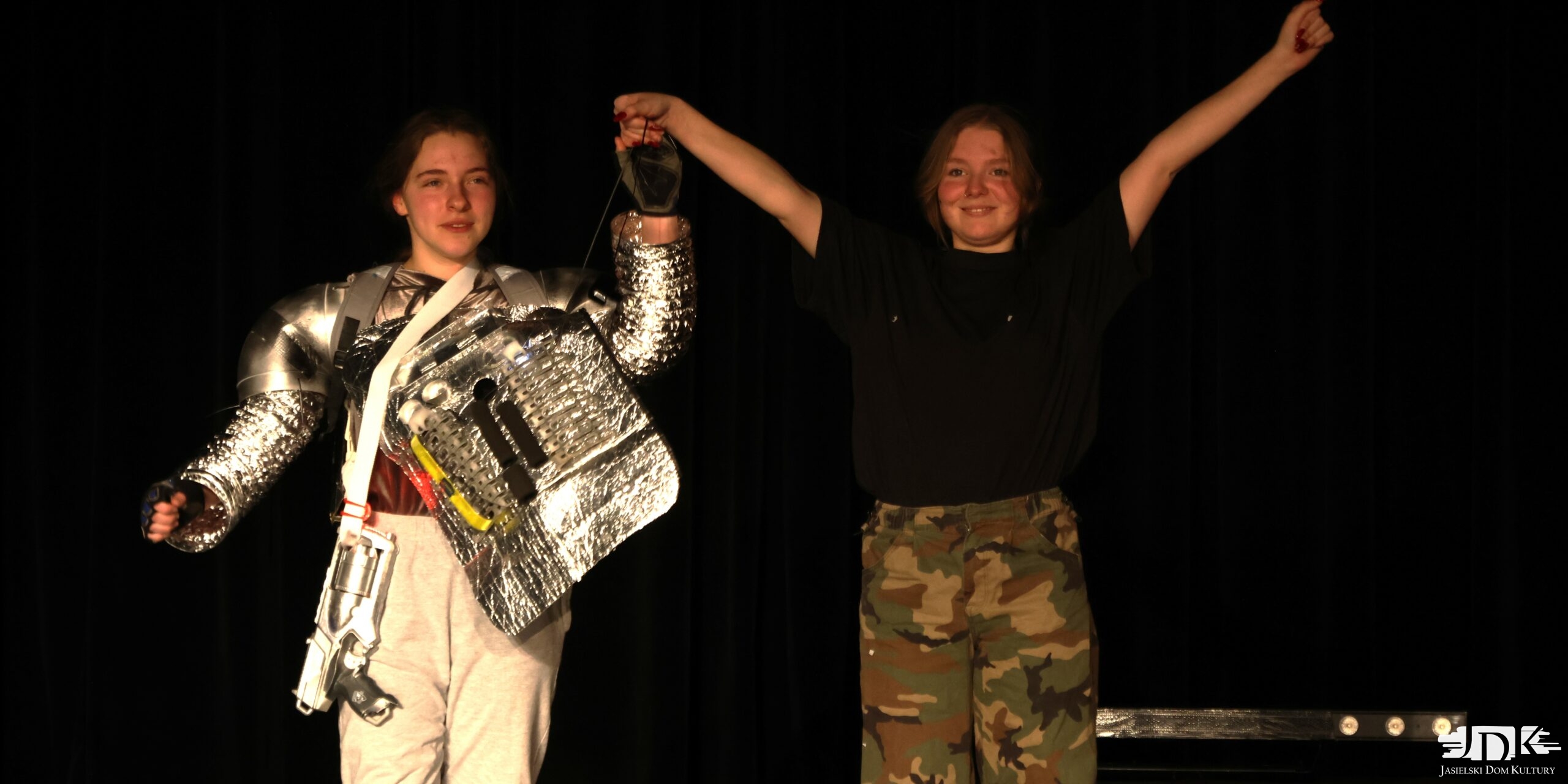 dziewczyna przebrana za robota - srebrny kostium z migającymi światełkami oraz dziewczyna w spodniach moro i czarnej koszulce, trzymające się za ręce podniesione ku górze