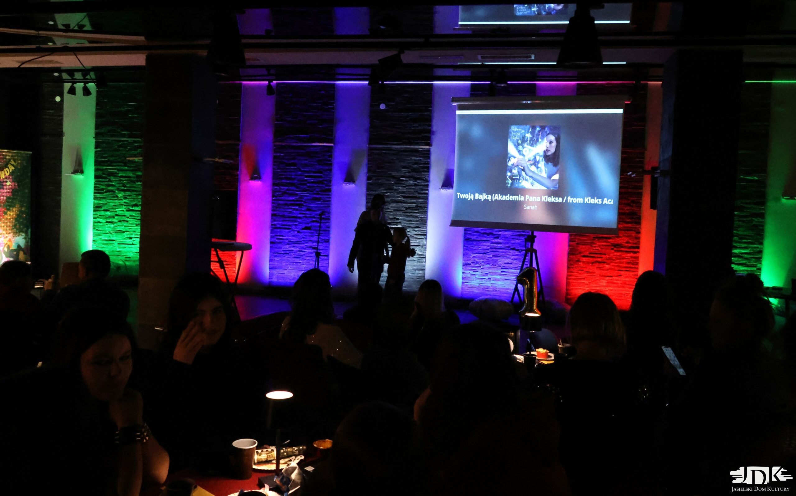 sala pełna ludzi z ekranem i wyświetlanym tekstem piosenki