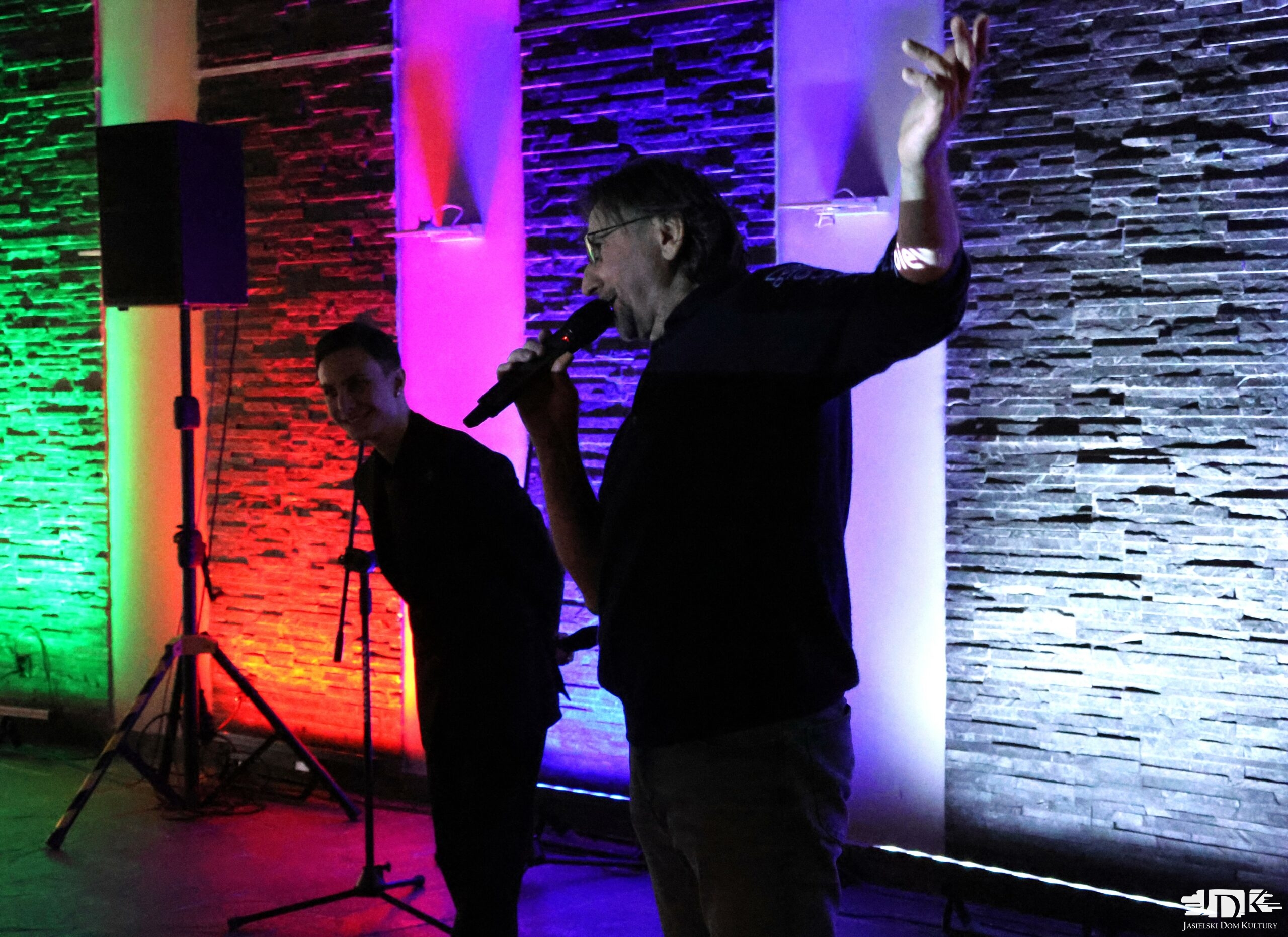 mężczyzna w okularach z wyciągniętą ręką, w tle drugi mężczyzna śmieje się do kamery