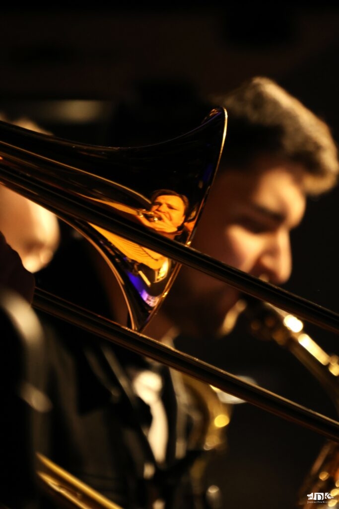 na pierwszym planie fragment puzonu z twarzą muzyka, w tle twarz saksofonisty