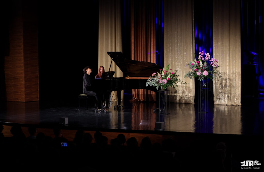 artysta przy fortepianie, obok kobieta, po drugiej stronie aranżacja kwiatowa