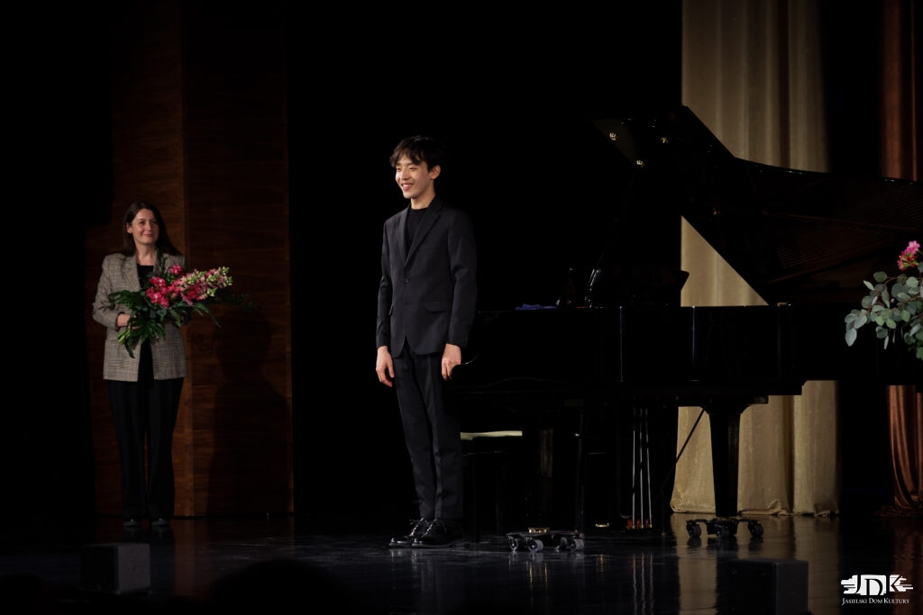 uśmiechnięty artysta stojący przy fortepianie, po jego prawej stronie dyrektorka JDK z kwiatami