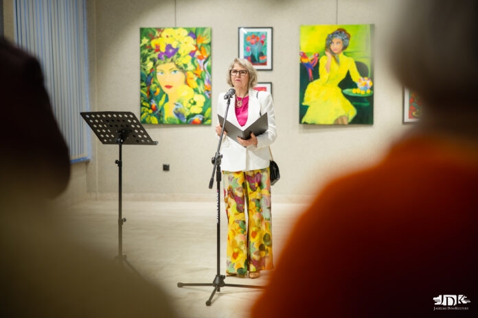 ikonka Wystawa Zofii Zmudy Hendriks, malarka stojąc na tle obrazów przemawia do publiczności