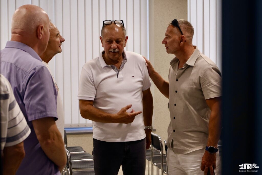 Grupa starszych mężczyzn rozmawia w pomieszczeniu z krzesłami, gestykulując i patrząc na siebie.