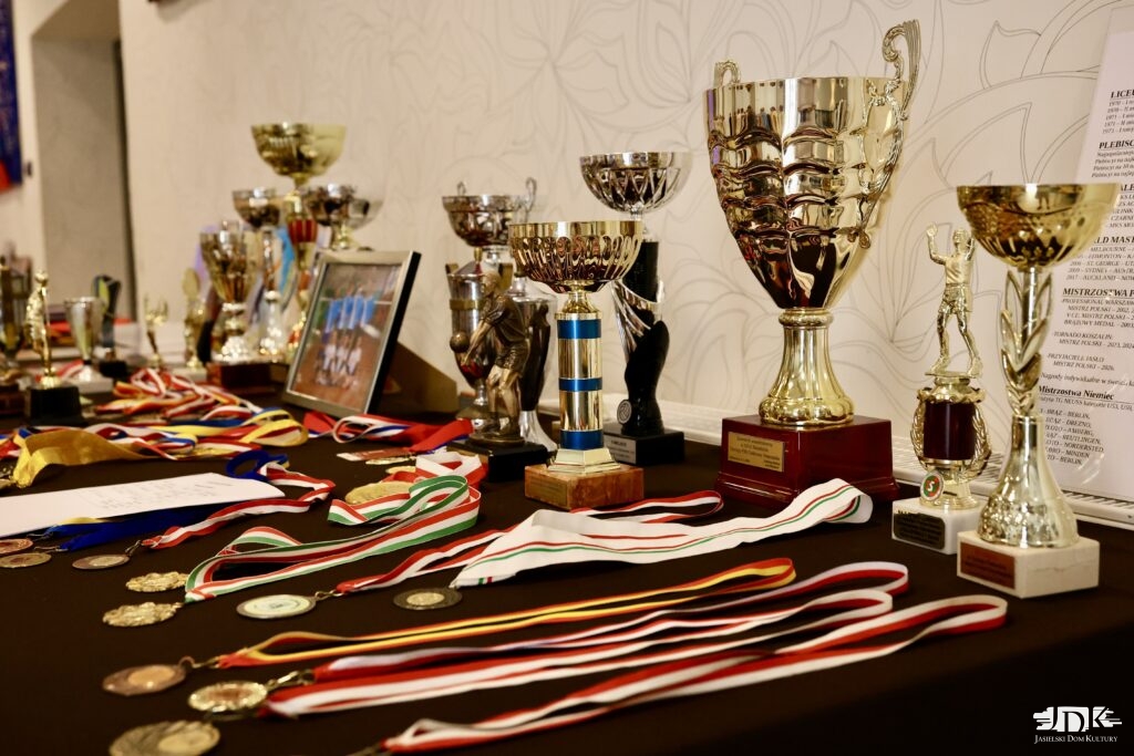 Stół z licznymi pucharami sportowymi, medalami na wstążkach oraz pamiątkowym zdjęciem w ramce.
