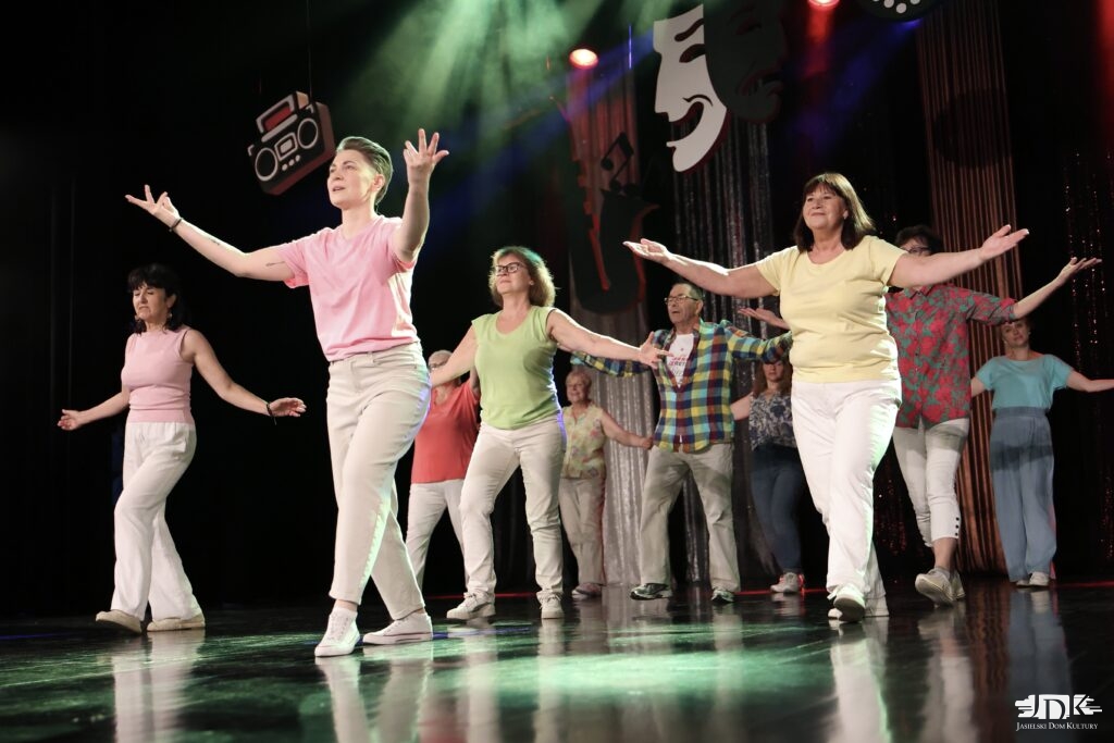 Grupa dorosłych w kolorowych koszulkach i jasnych spodniach tańczy na oświetlonej scenie teatralnej.