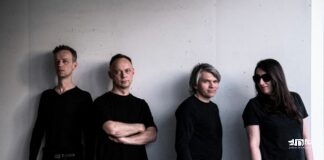 Cztery osoby w czarnych strojach pozują na tle jasnej, betonowej ściany w minimalistycznym otoczeniu.
