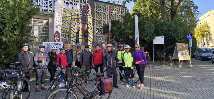 grupa trzynastu rowerzystów pod budynkiem JDK - w kaskach oraz sportowym ubraniu